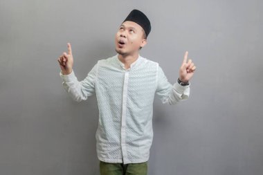 Mutlu Asyalı Müslüman adam tanıtımı fotokopi alanını işaret ederek oruç ayının gölgeleriyle koko gömlek ve göğüs kafesi giyerek gri bir arkaplanda izole edilmiş bir halkayı işaret ederek tavsiye ediyor.