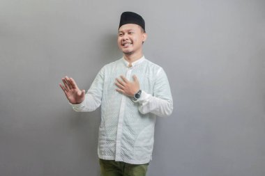 Asyalı Müslüman adam dur işareti yapıyor. Avuç içiyle Koko tişörtü ve oruç ayının gölgesinde peci giyiyor. Gri bir arka planda izole edilmiş.