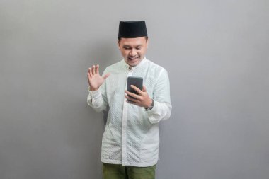 Asyalı Müslüman bir adam oruç ayının gölgesinde koko gömleği ve peki giyiyor, elini kaldırıyor ve akıllı telefonuna bakıyor, gri bir arka planda izole edilmiş.