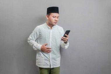 Asyalı Müslüman bir adam oruç ayının gölgeleriyle koko gömleği ve peki giyiyor, açlığı bastırıyor ve gri bir arkaplanda orucun kırılması için zamanı bekliyor.