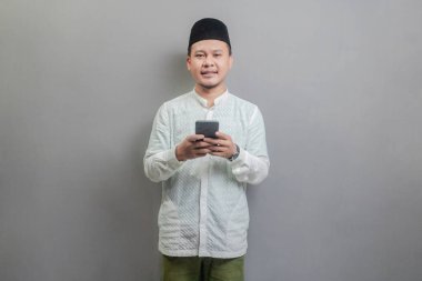 Mutlu Asyalı Müslüman adam aramalar için akıllı telefon kullanıyor, sosyal medya, mobil uygulama çevrimiçi oruç ayının gölgelerini taşıyan bir koko gömlek ve göğüs giyiyor, gri bir arka planda izole edilmiş.