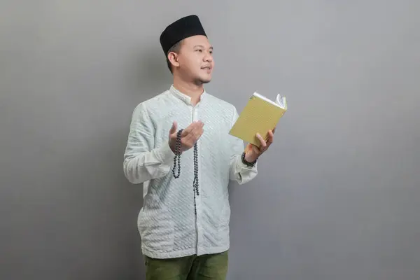 亚洲穆斯林男子 身穿可可可豆衬衫 头戴山核桃色的禁食月 手持祈祷珠子 古兰经 与灰色背景隔离 — 图库照片