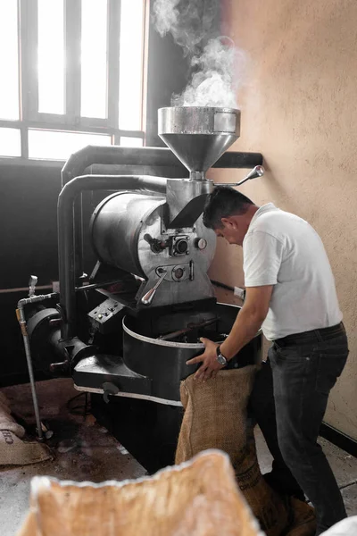 An Hispanic man is picking up the coffee with a sack from the roaster machine. Concept of local coffee production