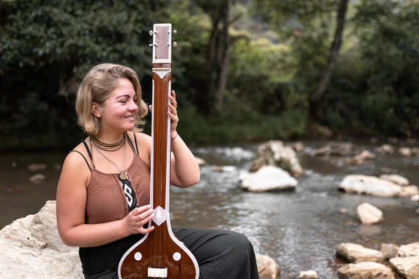 Genç yetişkin bir kadın nehir kayasında oturmuş Hint tanpurasını çalarken gülümsüyor. Açık havada müzik çalma kavramı
