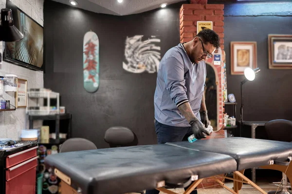 A tattoo artist is cleaning the studio stretcher with a spray. Concept of hygiene in the tattoo studio