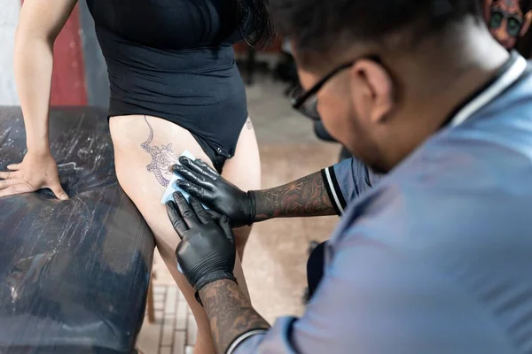 Dövme sanatçısı dövme yapmadan önce kadın bacağını temizler. Dövme stüdyosunda hijyen kavramı