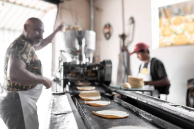 An adult man is working with his son producing corn tortillas with a nixtamall mill. Concept of traditional corn tortillas preparation clipart