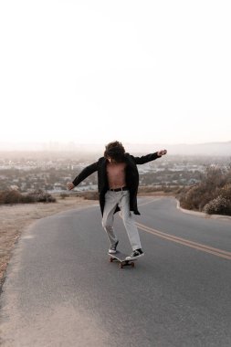 A shirtless young man wearing a gabardine is skating on a road in Baldwin Hills during the sunset, Los Angeles. Concept of skate lifestyle