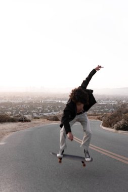 A shirtless young man wearing a gabardine is skating on a road in Baldwin Hills during the sunset, Los Angeles. Concept of skate lifestyle