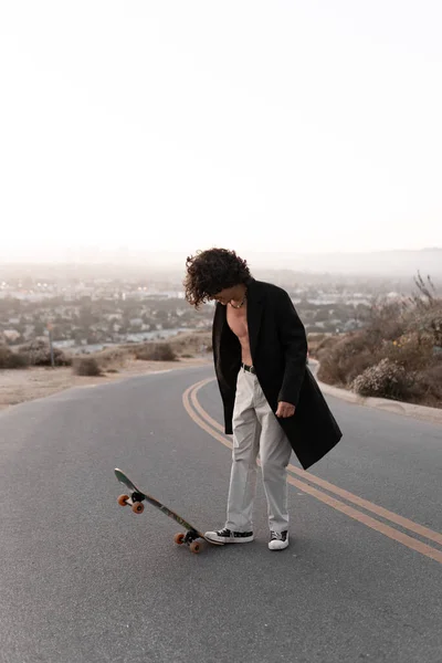 A shirtless young man wearing a gabardine is playing with his skate in Baldwin Hills during the sunset, Los Angeles. Concept of skate lifestyle
