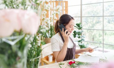 Beautiful Asian florist woman in apron talking on mobile phone  with clients or customers at flower shop, Female receiving order of flowers, Small business, entrepreneur concept