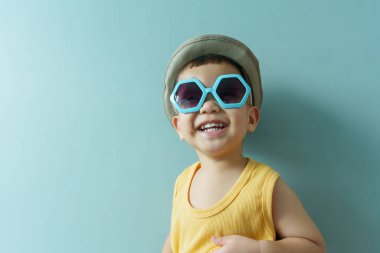 Portrait of Cheerful little Asian boy in yellow sleeveless and blue sunglasses on blue background. Smiling face