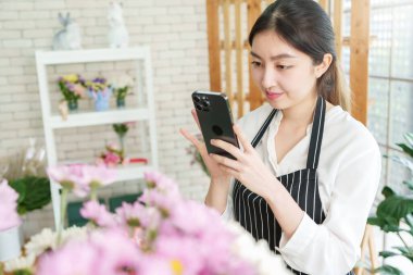 Güzel Asyalı kadın çiçekçi çiçekçi çiçekçide akıllı telefonlu çiçeklerin fotoğrafını çekiyor, küçük işletme sahibi, girişimci, cep telefonu kullanıyor.