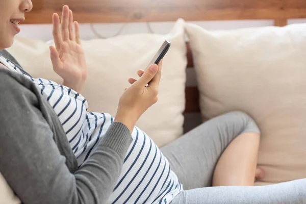ソファーに座っているスマートフォンにビデオ通話と手を振っているアジアの妊婦 幸せな若いですお母さんと話して彼女の夫で携帯電話で自宅 — ストック写真