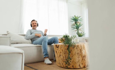 Sakallı yetişkin bir adam kanepeye oturmuş kulaklıkla müzik dinliyor ve gitar çalıyor, yaşam tarzı kavramı, evde rahatlama..