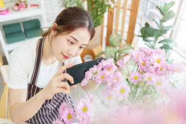 Güzel Asyalı kadın çiçekçi çiçekçi çiçekçide akıllı telefonlu çiçeklerin fotoğrafını çekiyor, küçük işletme sahibi, girişimci, cep telefonu kullanıyor.