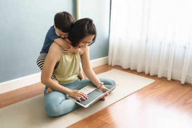 Spor giyimli sağlıklı bir Asyalı kadın oğlunu taşırken yoga minderinde dizüstü bilgisayar kullanıyor. Anne oğluyla binicilik oynuyor. Annelik