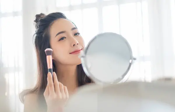 Beauty portrait of Happy smiling Asian Korean Female model using makeup brush and looking at makeup mirror at home. Skin care, Hairstyle.