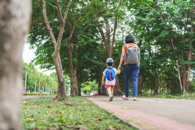 Arka manzara, sırt çantalı ve şapkalı küçük Asyalı çocuk anaokuluna giderken annesinin elini tutuyor. Okula geri dön..