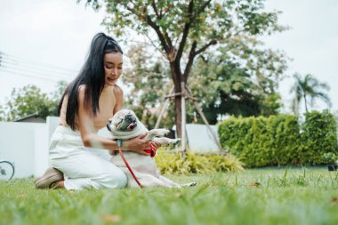 Neşeli Asyalı Kadın Pug köpeğini Sunny Garden 'daki çimenlikte gıdıklıyor.