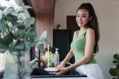 Canlı yeşil kolsuz gülümseyen Asyalı genç kadının portresi güneşli, modern bir mutfakta tabak temizliyor.