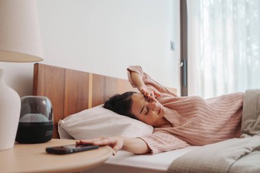 Yumuşak pembe kazaklı genç bir Asyalı kadın baş ağrısı ile yatakta yatıyor, elleri alnında, sakin bir ifade ve pencereden içeri akan doğal ışıkla..