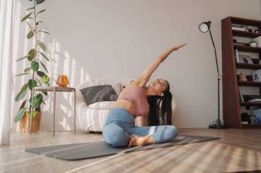 Oturan Asyalı kadın yoga pozisyonu almış, kollarını sıcacık, güneşli bir yaşam alanında uzatıyor.