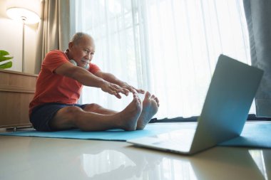 Evde yoga yapan yaşlı Asyalı bir adam, bir dizüstü bilgisayardaki online dersi takip ediyor.
