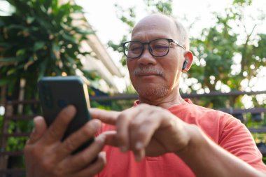 Bahçe 'de akıllı telefon ve kulaklık kullanan kıdemli Asyalı adam dışarıda teknolojiyle meşgul ve mutlu.