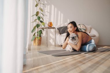 Sevecen genç Asyalı kadın pug köpeğini yoga minderinde kucaklıyor güneşli odada hassas bir anı paylaşıyor.