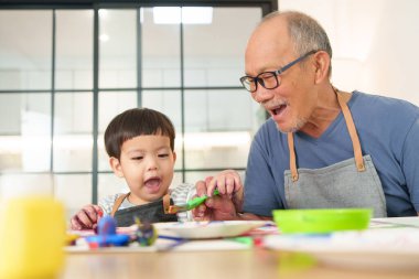 Asyalı son sınıf öğrencisi ve küçük çocuk ya da torun evde neşeli ve yaratıcı aile ilişkilerini vurgulayarak bir masada birlikte resim yapmaktan zevk alıyorlar..