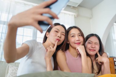 Parlak bir odada cep telefonuyla selfie çeken üç genç Asyalı kadın eğlenceli ve neşeli ifadeler sergiliyorlar..