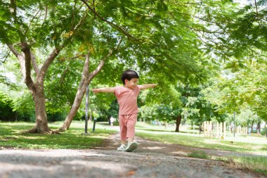 Pembe elbiseli Asyalı küçük çocuk parkta ağaçlık bir patika boyunca mutlu bir şekilde koşuyor, kolları açık, güneşli bir günde yandan yakalanmış..