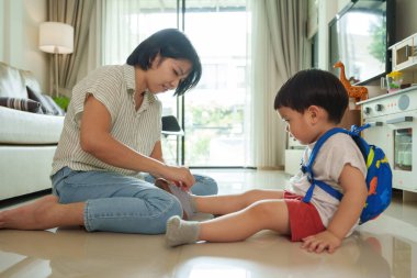 Asyalı anne çocuk ya da oğulun ayakkabılarını giymesine yardım ediyor, evde dışarı çıkmaya hazırlanıyor, aydınlık ve güneşli iç mekan, sabah rutini.