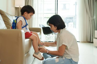 Asyalı anne, küçük oğlunun oturma odasında ayakkabı giymesine yardım ediyor. Anne bağcıklarını bağlarken sırt çantalı çocuk kanepede oturuyor. Sabah rutini, ebeveyn-çocuk bağı, okul hazırlığı..
