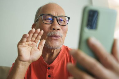 Gözlüklü ve turuncu tişörtlü Asyalı yaşlı bir adam akıllı telefona bakarken el sallıyor, koltukta mutlu bir ifadeyle oturuyor, ailesiyle video görüşmesi yapıyor.