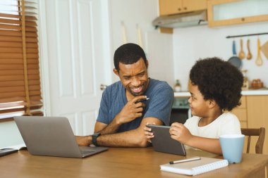 Siyah Afrika kökenli Amerikalı baba ve çocuk evdeki mutfak masasında tablet ve laptop kullanıyorlar. Sıradan bir ev hayatı. Aile bağları ve evde öğrenim kavramı.