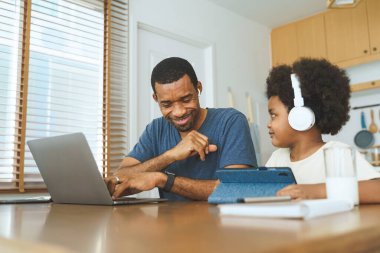Siyah Afrikalı Amerikalı baba ve çocuk ev mutfak masasında dizüstü bilgisayar ve kulaklık kullanıyor. Sıradan bir ev hayatı. Aile bağları ve evde öğrenim kavramı.