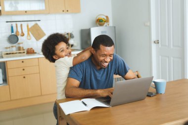 Siyah Afro-Amerikan Baba dizüstü bilgisayarda çalışırken çocuk evde mutfak masasında ona şakayla sarılıyor. Sıradan bir ev hayatı. Aile bağı ve uzaktan çalışma kavramı.