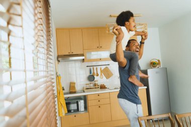 Afro-Amerikan babası genç oğlunu mutlu bir şekilde omuzlarında taşıyarak, hem gülüyor hem de gülümsüyor, içten ve neşeli bir aile bağı ve mutluluk anı yaratıyor..
