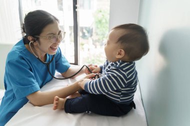 Parlak sağlık muayenehanesinde bebeğe gülümseyen Asyalı çocuk doktoru, hastanede steteskoplu bir çocuğu muayene eden doktor..