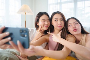 Üç genç Asyalı kadın bir kanepede yan yana oturuyor. Parlak bir oturma odasında akıllı bir telefonla selfie çekerken neşeli yüzler ve jestler yapıyorlar..