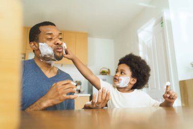 Siyah Afrikalı Amerikalı Baba ve küçük oğlu mutfak ortamında tıraş köpüğü sürüyorlar. Kapalı alan fotoğrafçılığı. Aile bağları ve bakım kavramı.