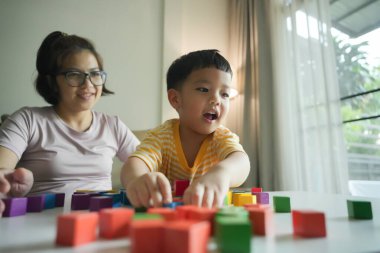 Asyalı anne gülümserken küçük oğlu renkli tahta bloklarla oynuyor, eğitici ve eğlenceli aktiviteler yapıyor, kapalı mekan ışığı, gündelik ortam, bağ kurma anı..