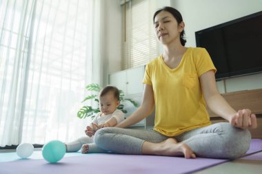 Asyalı bir kadın ve bir bebek bir odada paspasın üzerinde oturuyorlar. Kadın sarı bir gömlek giyiyor ve bebeğin elinde mavi bir top var. Kadın olarak sakinlik ve rahatlama kavramı.