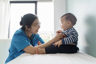 Mavi önlüklü Asyalı kadın hemşire, klinikte steteskoplu meraklı bir çocuğu muayene ediyor, bebeklerle iletişim kuran gülümseyen bir çocuk doktoru...