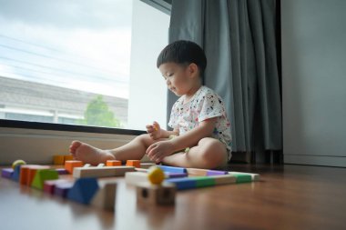 Asyalı genç çocuk pencerenin kenarında oturmuş renkli tahta bloklarla oynuyor..