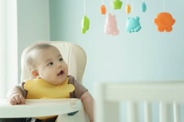 Asyalı gülümseyen bebek sandalyesinde asılı oyuncaklara bakıyor, nişanlı ve mutlu, erken çocukluk.