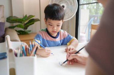Asyalı genç bir çocuk, annesiyle birlikte renkli bir kalemle bir kağıt çiziyor..