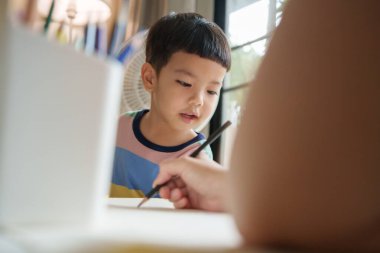 Mutlu Asyalı çocuk bir masada oturuyor ve kağıt üzerinde renkli kalemle boyuyor. Yaratıcılık ve oyunculuk kavramı